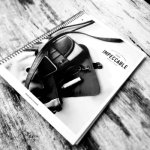 Spiral-bound notebook titled "Impeccable" rests on a wooden surface with various accessories like sunglasses and a small bag.